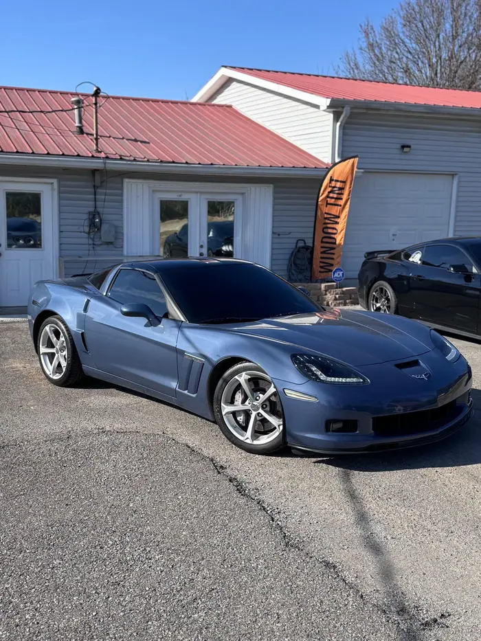 Blue Corvette Grand Sport with dark window tint outside the Stealth Tint and PPF shop on Highway 109, a Window Tint flag beside the door