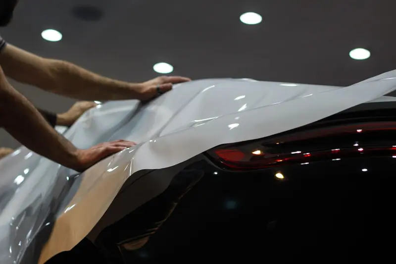 Two hands laying vinyl film over a rear quarter panel under shop lights (stock photo)